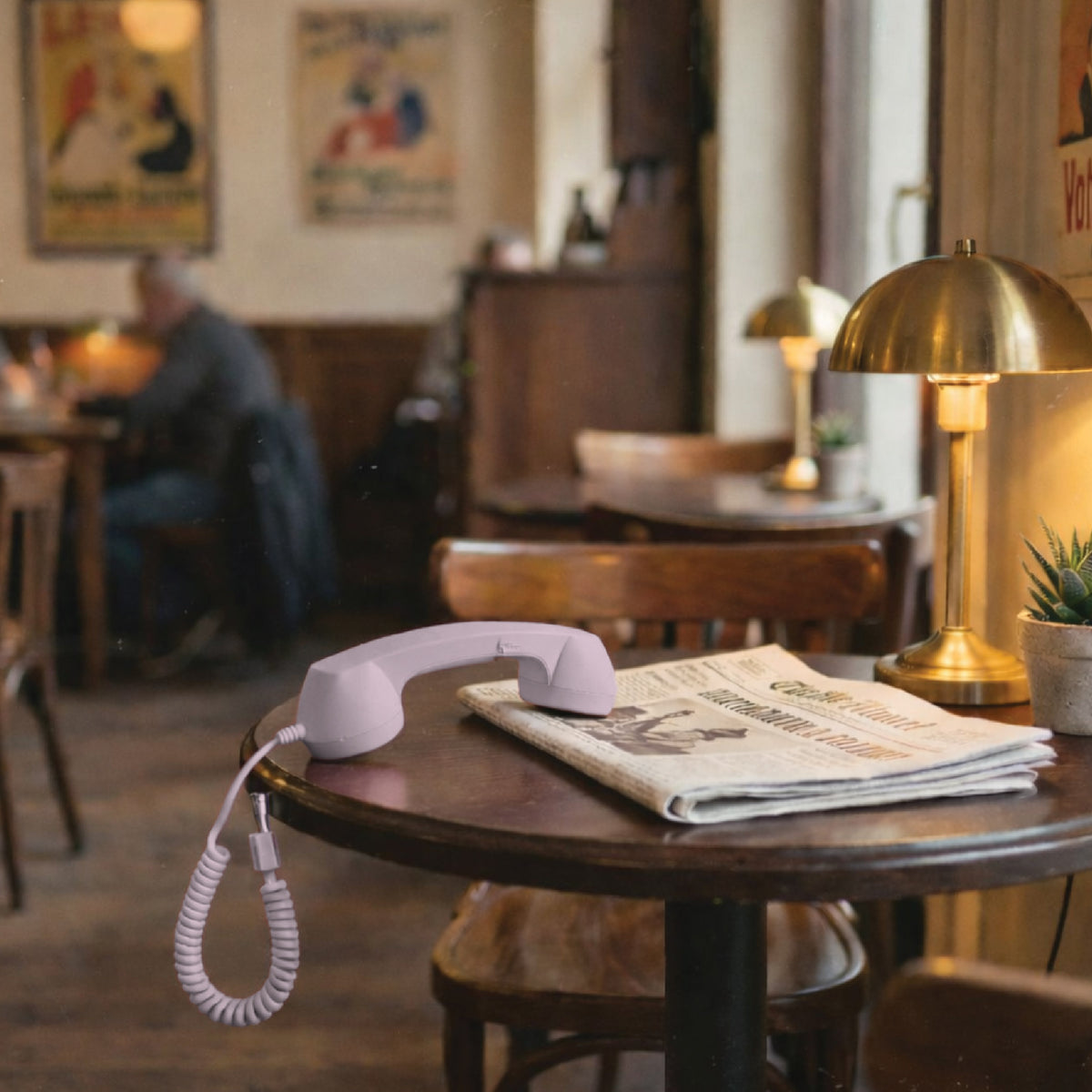 Pink USB-C retro phone handset on a café table beside a newspaper, highlighting a vintage-inspired accessory for everyday calls on the go